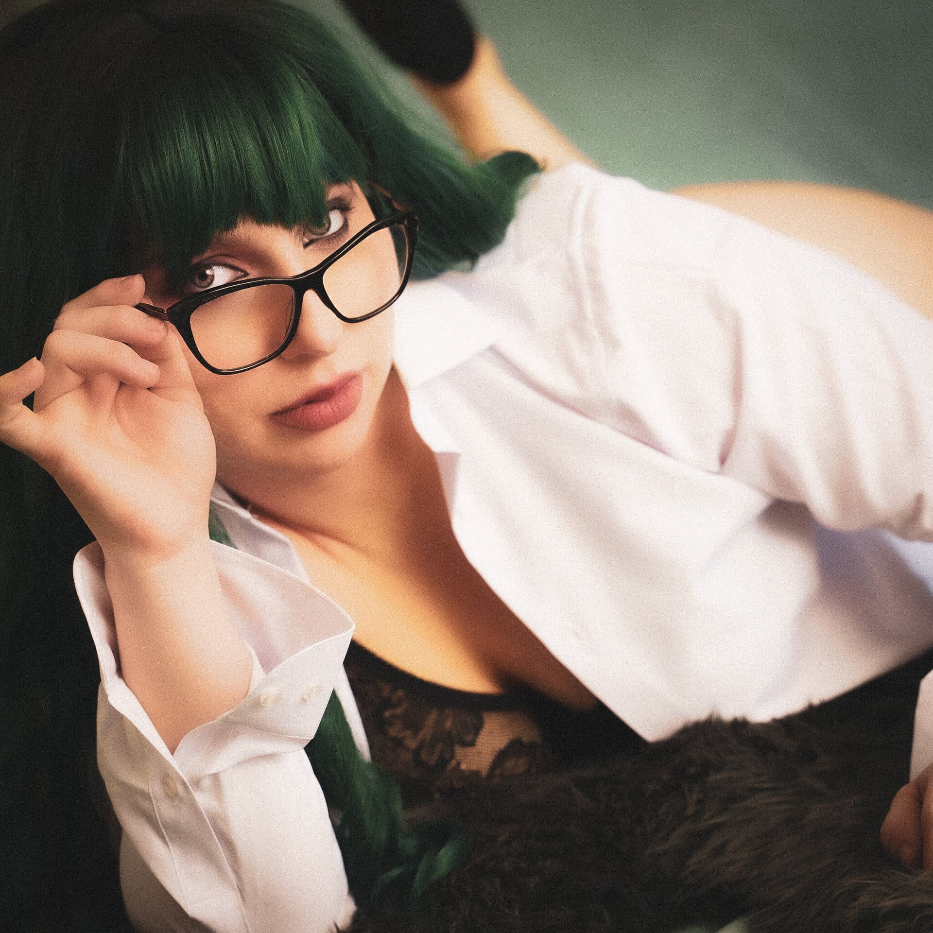 A person wearing a green wig and glasses lying down, adjusting their glasses while gazing towards the viewer. they are dressed in a white shirt layered over a lace top. Image by Rya Eisma of Shamelessly Sexy Boudoir Photography, Las Vegas, NV