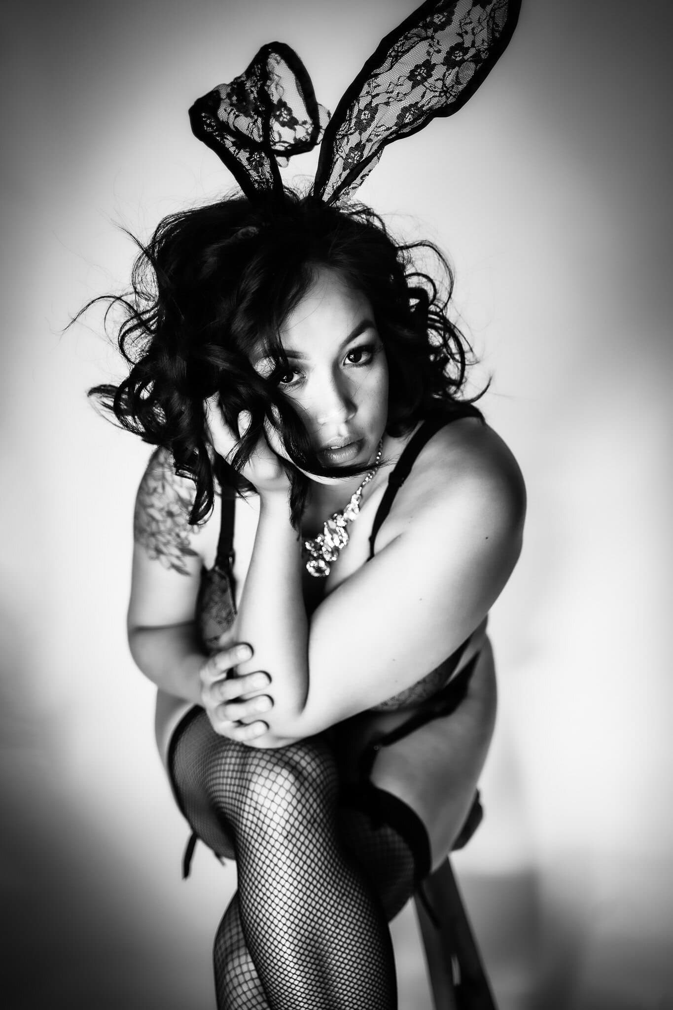 A woman in lingerie, fishnet stockings, and lace bunny ears poses sitting on a stool. She has curly hair, a tattoo on her upper arm, and gazes intensely at the camera. The image is black and white. Image by Rya of Shamelessly Sexy Boudoir Photography, Las Vegas, NV