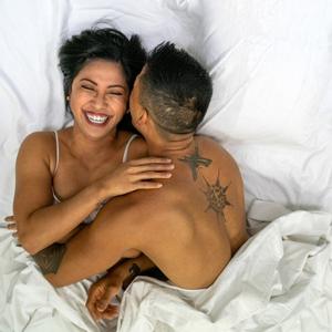 couples boudoir. asian couple, both brunette, and tan/brown skinned, are in bed laughing, lying on white sheets, covered up to the waist by white comforter. Male has tattoos on his arms and back, female is wearing a white camisole tank. Male is turned on his side to hug female, female's hand is on his shoulder.