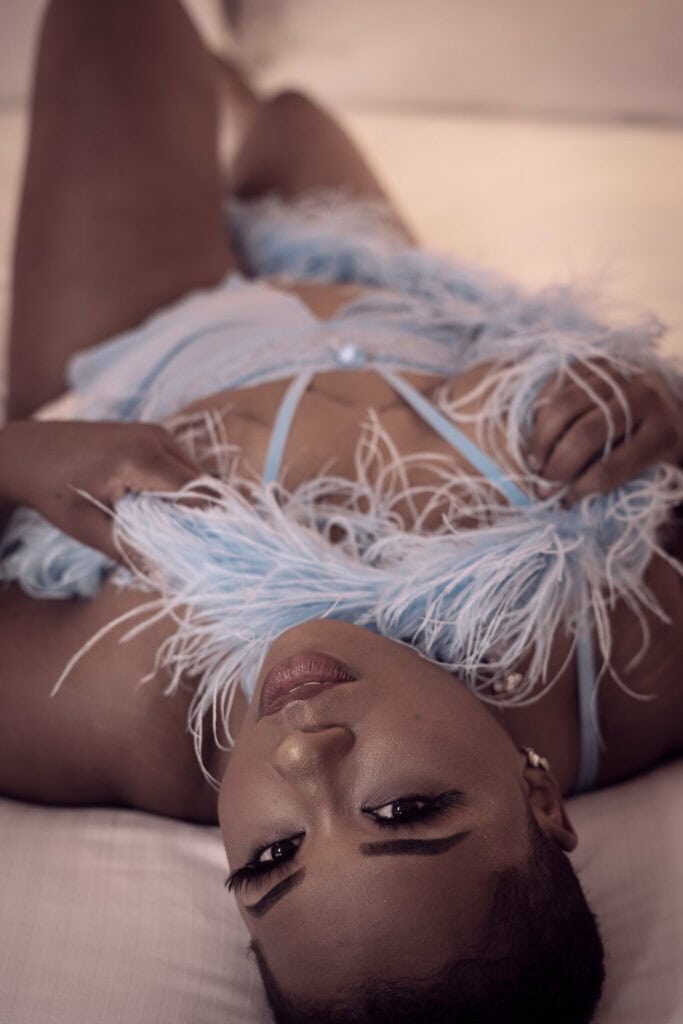 A woman wearing a light blue feathered lingerie lies gracefully on her back, her gaze fixed upwards, highlighting her expressive eyes and striking makeup. Image by Rya Eisma of Shamelessly Sexy Boudoir Photography, Las Vegas, NV