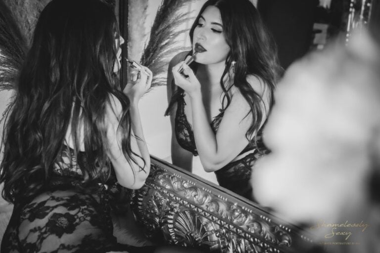 black & white image of beautiful long haired brunette woman in black lace leaning in and applying lipstick in mirror reflection as form of self-care