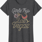 Heather gray T-shirt bedazzled with "Girl's Trip" in clear/silver rhinestones, then dice and a tiled glass of red wine, then in gold rhinestones it reads "Las Vegas".