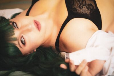 A woman with green hair and red lipstick lies on a bed, gazing upward. she is wearing a black lace garment and partially covered by a white sheet. Image by Rya Eisma of Shamelessly Sexy Boudoir Photography, Las Vegas, NV