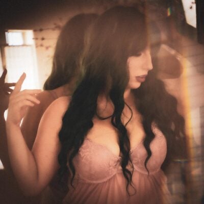 A woman in a soft pink dress, reflected in a mirror as she adjusts her long, wavy black hair. the lighting creates a warm, nostalgic aura. Image by Rya Eisma of Shamelessly Sexy Boudoir Photography, Las Vegas, NV