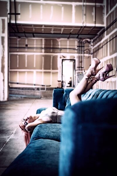 A woman with pink hair lying on a blue velvet couch in an industrial-style building with exposed piping on the ceiling. she wears a light outfit and high heels, relaxing with her feet elevated. Image by Rya Eisma of Shamelessly Sexy Boudoir Photography, Las Vegas, NV