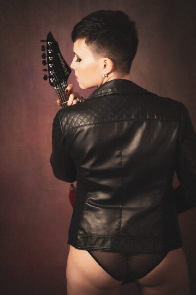 A person with a short haircut and wearing a leather jacket, viewed from the back, holds an electric guitar over their shoulder. a warm, moody background enhances the artistic vibe. Image by Rya Eisma of Shamelessly Sexy Boudoir Photography, Las Vegas, NV