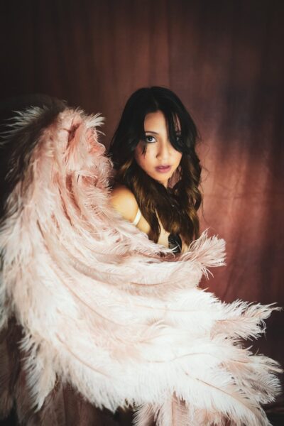 A woman with long wavy hair looks over her shoulder, wrapped in a soft, pink feathered cloak against a dark red backdrop. her expression is engaging and mysterious. Image by Rya Eisma of Shamelessly Sexy Boudoir Photography, Las Vegas, NV