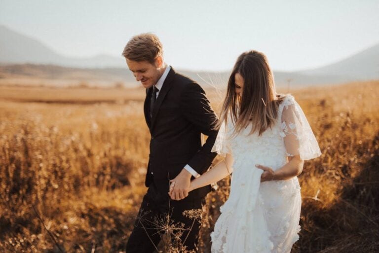 A young couple, dressed in a suit and a white lace dress, walk hand in hand through a sunlit field with mountains in the background. Image by Rya Eisma of Shamelessly Sexy Boudoir Photography, Las Vegas, NV