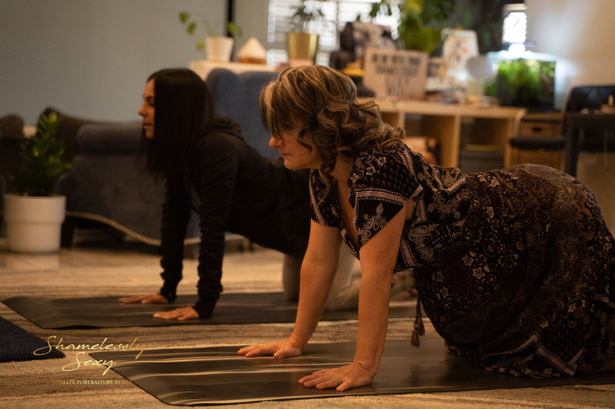 Two women practicing yoga indoors, both in a cow pose with hands and knees on the floor. They appear focused and relaxed in a warmly lit room with comfortable furniture and potted plants. The background shows a sign that reads "Shamelessly Sexy. Image by Rya Eisma of Shamelessly Sexy Boudoir Photography, Las Vegas, NV