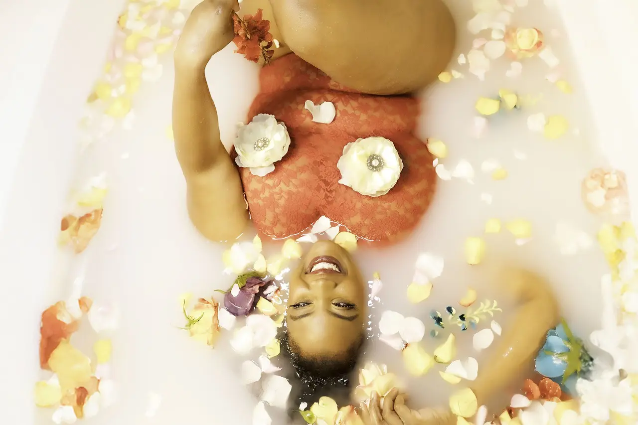 A person relaxes in a milky bath with scattered petals. They wear a red, lacy top adorned with white flower pasties, and colorful flower petals are spread around them. The person is smiling, lying on their back with one leg bent. Image by Rya Eisma of Shamelessly Sexy Boudoir Photography, Las Vegas, NV