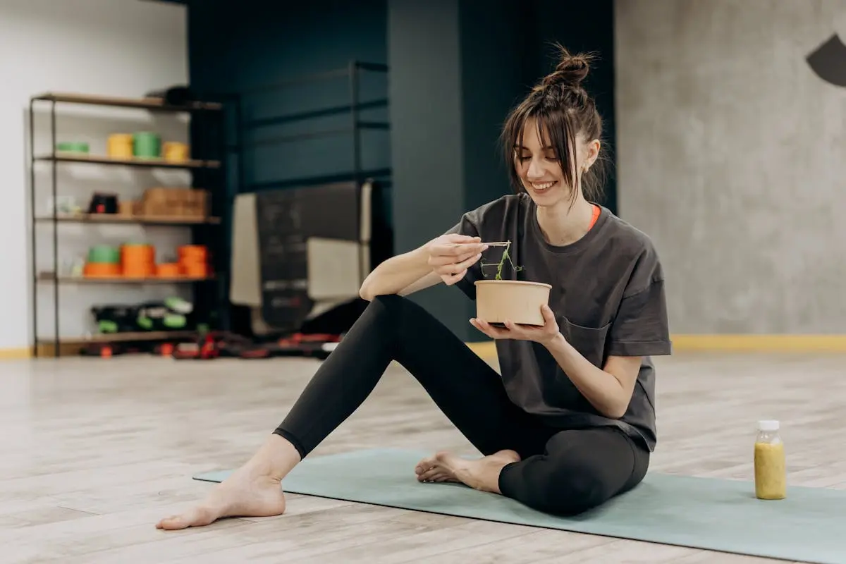 A woman with brown hair in a bun sits on a yoga mat in a gym, smiling and eating from a bowl with chopsticks. She is wearing a gray t-shirt and black leggings. A yellow drink in a bottle is placed on the mat beside her. Equipment is visible in the background. Image by Rya Eisma of Shamelessly Sexy Boudoir Photography, Las Vegas, NV