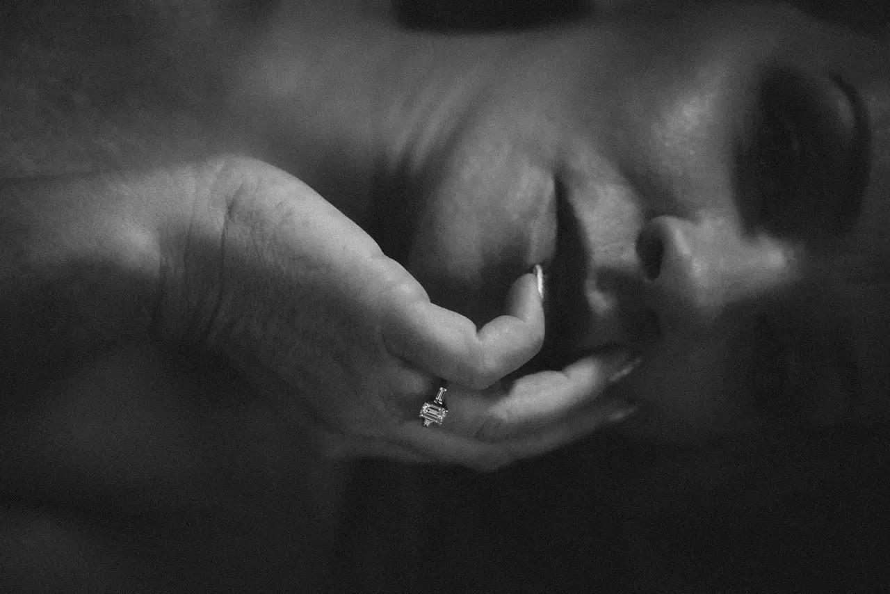A grayscale close-up image of a person's face and hand. The hand, adorned with a ring featuring a rectangular gemstone, gently touches their parted lips. The person's expression appears contemplative, with soft lighting creating shadows on their face. Image by Rya Eisma of Shamelessly Sexy Boudoir Photography, Las Vegas, NV