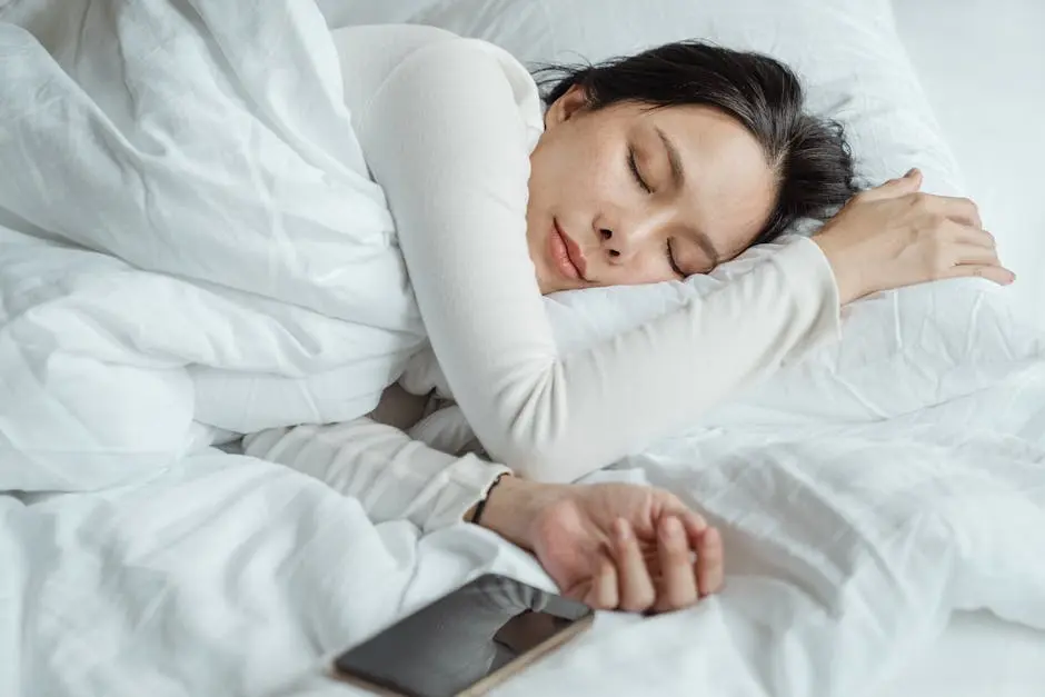 A person sleeping peacefully in bed, covered with a white blanket. Their head rests on a white pillow, and their arm is stretched out beside them. A smartphone lies next to their hand on the bed. Image by Rya Eisma of Shamelessly Sexy Boudoir Photography, Las Vegas, NV