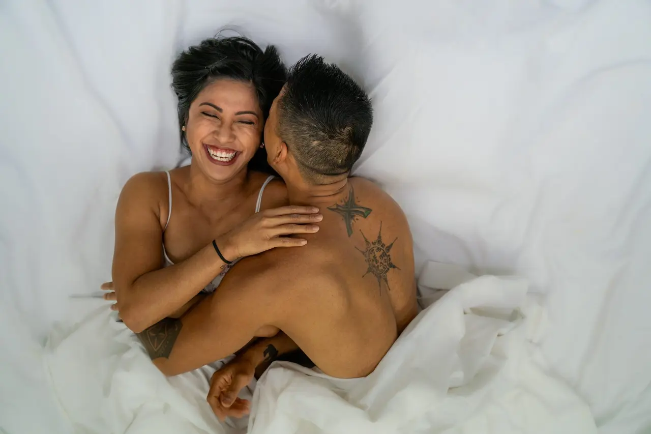 A couple lying on white sheets, joyfully embracing. The woman is smiling with her eyes closed, while the man has his back to the camera, revealing star tattoos on his back. They appear relaxed and happy. Image by Rya Eisma of Shamelessly Sexy Boudoir Photography, Las Vegas, NV