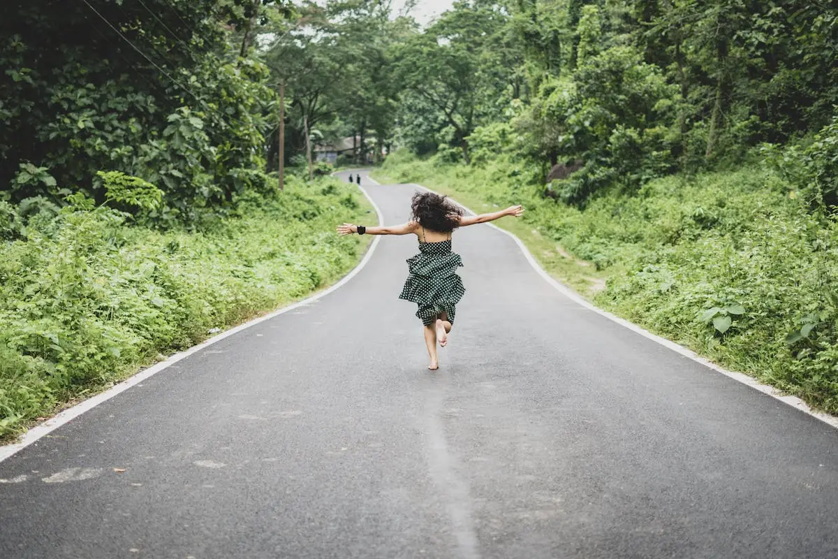 A woman in a black polka dot dress joyfully runs barefoot down an empty rural road surrounded by lush green trees on both sides. Her arms are spread wide, and her curly hair flows in the wind. The scene conveys a sense of freedom and adventure. Image by Rya Eisma of Shamelessly Sexy Boudoir Photography, Las Vegas, NV