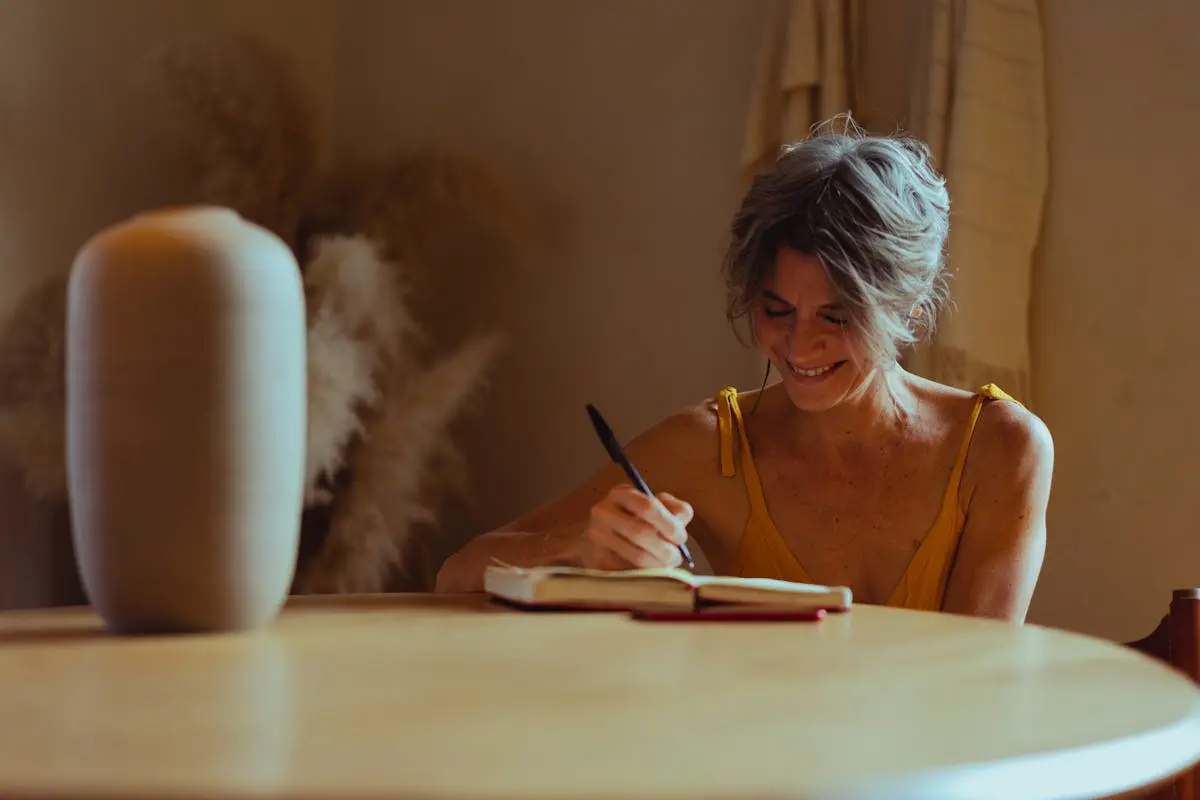 A woman with short hair sits at a round wooden table, writing in a notebook. She is wearing a yellow top and smiling. A large beige vase is on the table, and dried pampas grass is visible in the background. The setting is warm and cozy. Image by Rya Eisma of Shamelessly Sexy Boudoir Photography, Las Vegas, NV