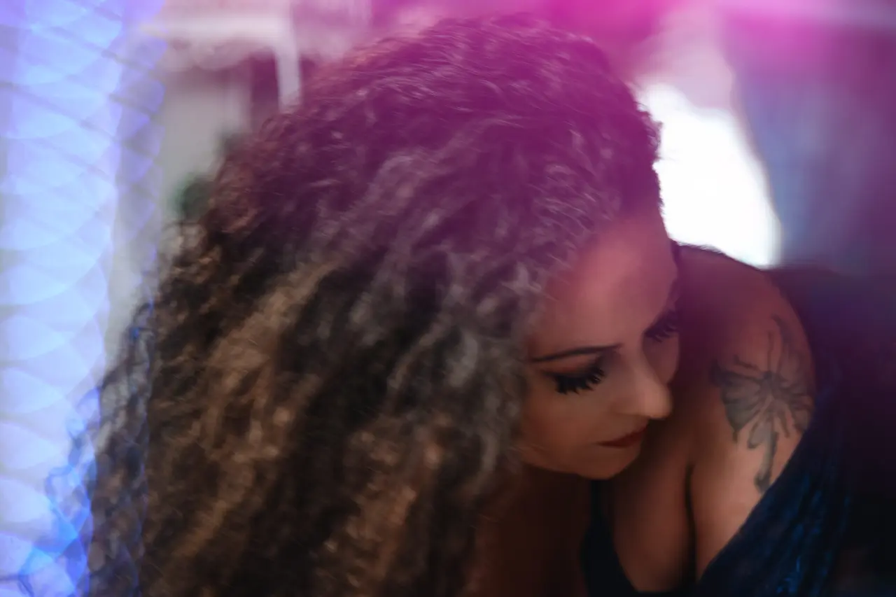 A woman with long curly hair looks downward. She has a tattoo of a flower on her shoulder. The image is softly lit with a pink and purple glow. Image by Rya Eisma of Shamelessly Sexy Boudoir Photography, Las Vegas, NV