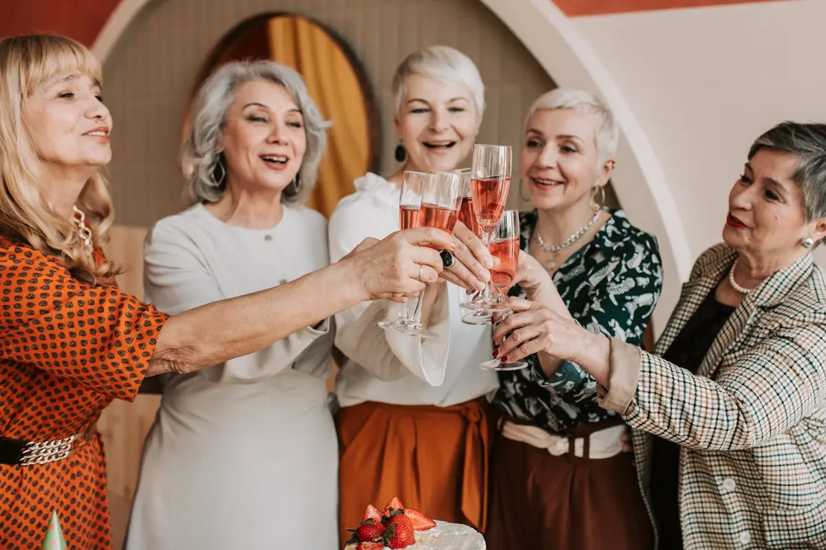 A group of five older women smiling and holding glasses of rosé champagne, making a toast around a table with a cake topped with strawberries. They are indoors, dressed in smart-casual attire, enjoying a cheerful celebration. Image by Rya Eisma of Shamelessly Sexy Boudoir Photography, Las Vegas, NV