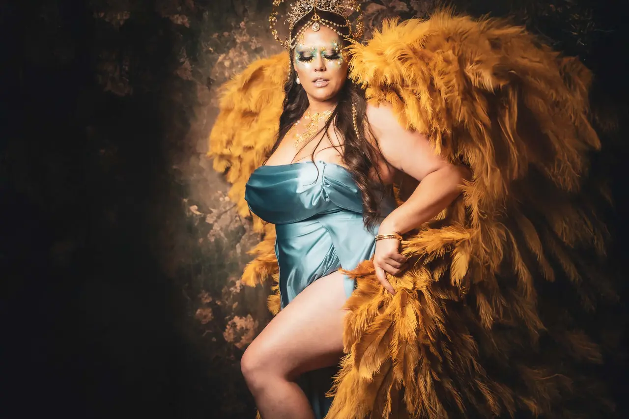 A person poses confidently wearing a teal satin dress and large golden feathered wings. They have an elaborate headpiece and bold makeup. The background is dark and textured, enhancing the dramatic effect of the costume. Image by Rya Eisma of Shamelessly Sexy Boudoir Photography, Las Vegas, NV