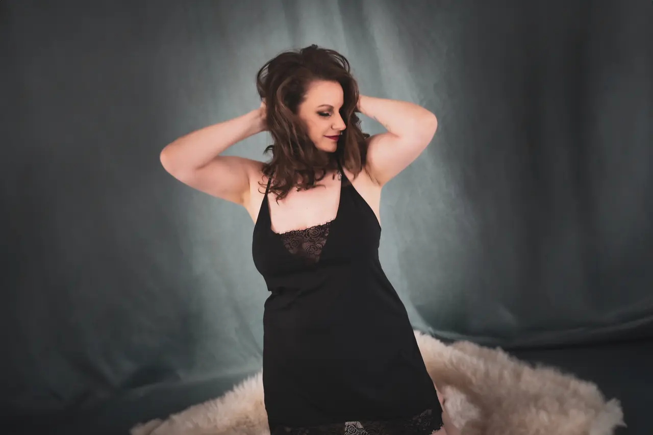 A woman in a black lace nightgown stands on a fluffy white rug, posing with her hands in her hair and looking to the side against a dark, softly lit backdrop. Image by Rya Eisma of Shamelessly Sexy Boudoir Photography, Las Vegas, NV
