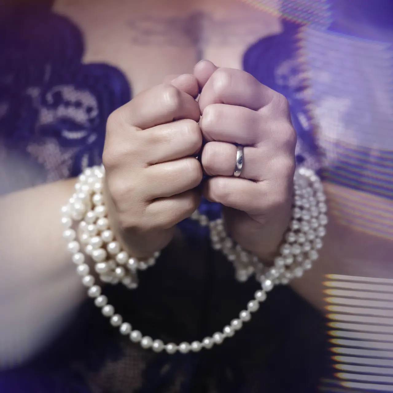 Close-up of two hands bound together with a string of pearls. One hand wears a ring. The person is dressed in dark, lacy clothing, and the background is softly blurred with light effects. Image by Rya Eisma of Shamelessly Sexy Boudoir Photography, Las Vegas, NV