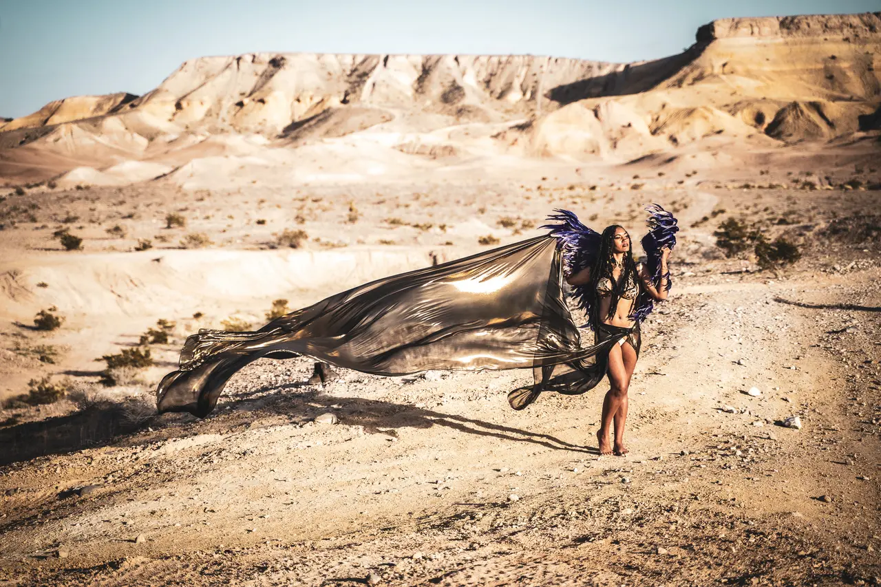 A person stands in a desert landscape wearing a shiny, flowing cape and dark wings. The backdrop features rocky hills bathed in sunlight, casting long shadows on the arid ground. Image by Rya Eisma of Shamelessly Sexy Boudoir Photography, Las Vegas, NV